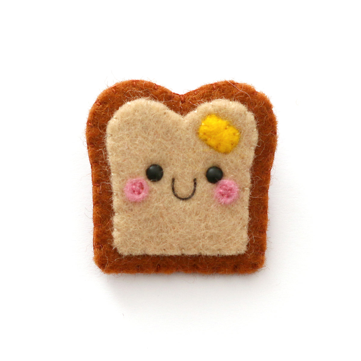 A cute, handcrafted felt pin shaped like a slice of toast with a smiling face, featuring a yellow butter detail, pink cheeks, and black eyes, set against a white background.