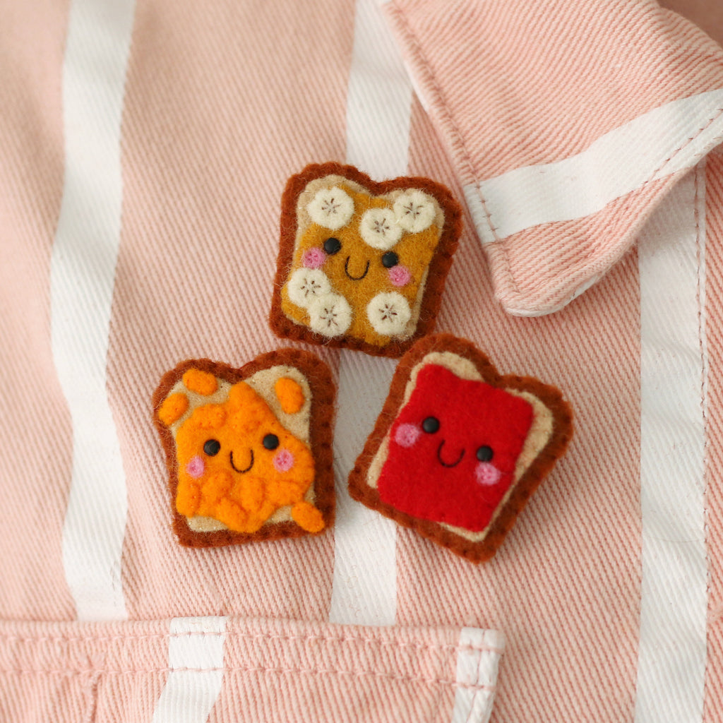 Three cute felt toast-shaped accessories with smiling faces are arranged on a pink and white striped jacket.