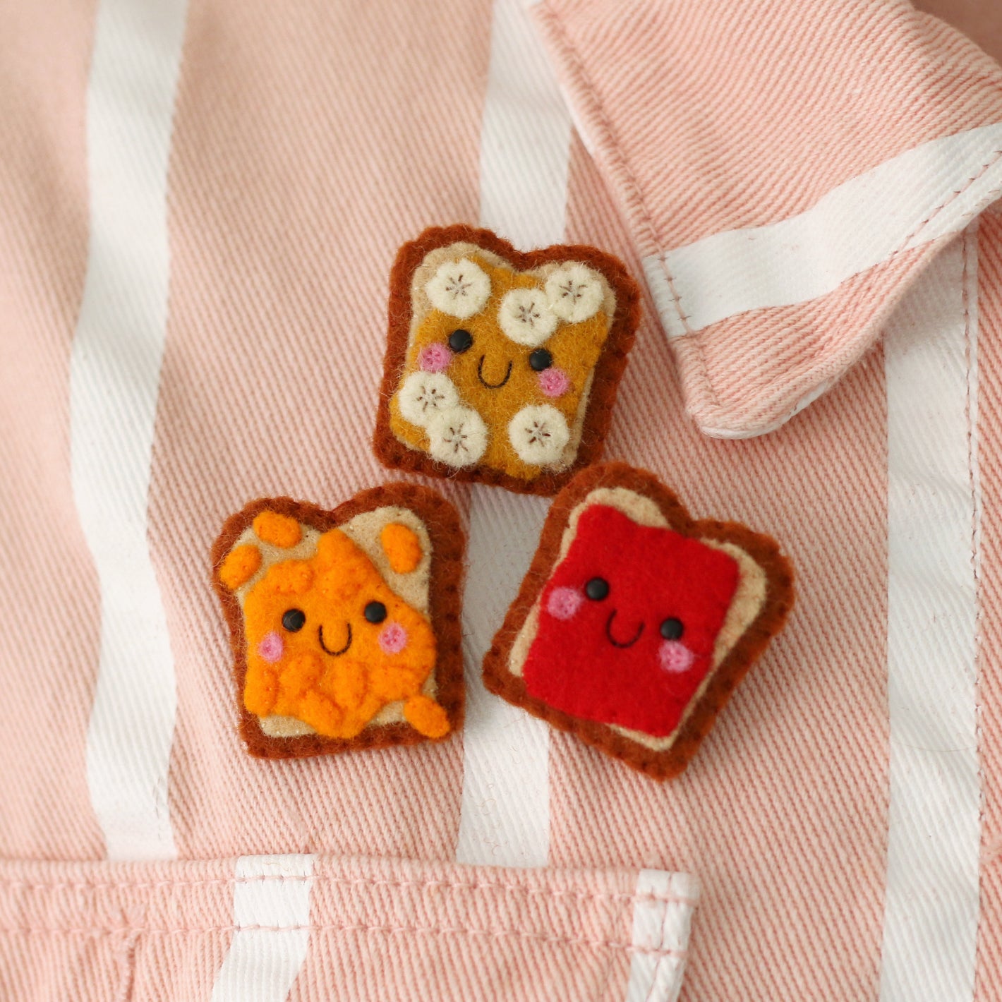 Three cute felt toast-shaped accessories with smiling faces are arranged on a pink and white striped jacket.