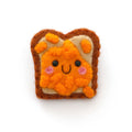 A cute felt pin shaped like a slice of toast with orange baked beans and a smiling face, on a plain white background.