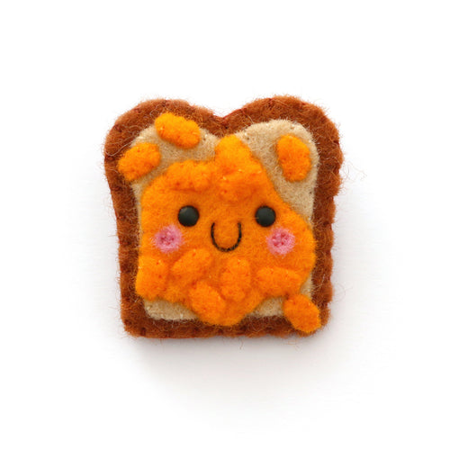 A cute felt pin shaped like a slice of toast with orange baked beans and a smiling face, on a plain white background.