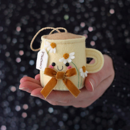 Small felt camomile tea mug with flowers and a bow held in a hand against a blue sequin background
