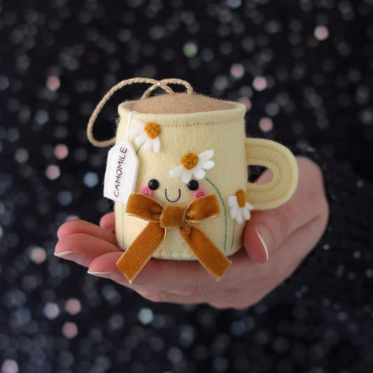 Small felt camomile tea mug with flowers and a bow held in a hand against a blue sequin background