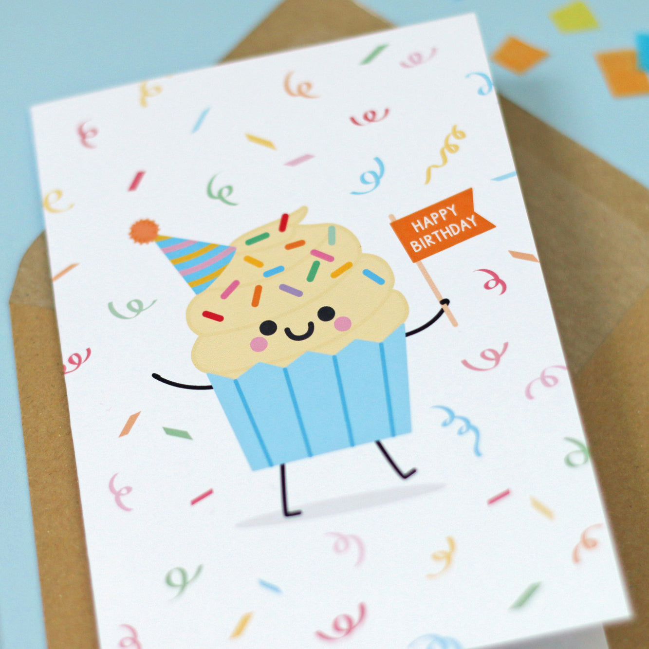 close up of kawaii style greeting card, featuring a vanilla cupcake with party hat and orange banner with the words 'HAPPY BIRTHDAY' on it