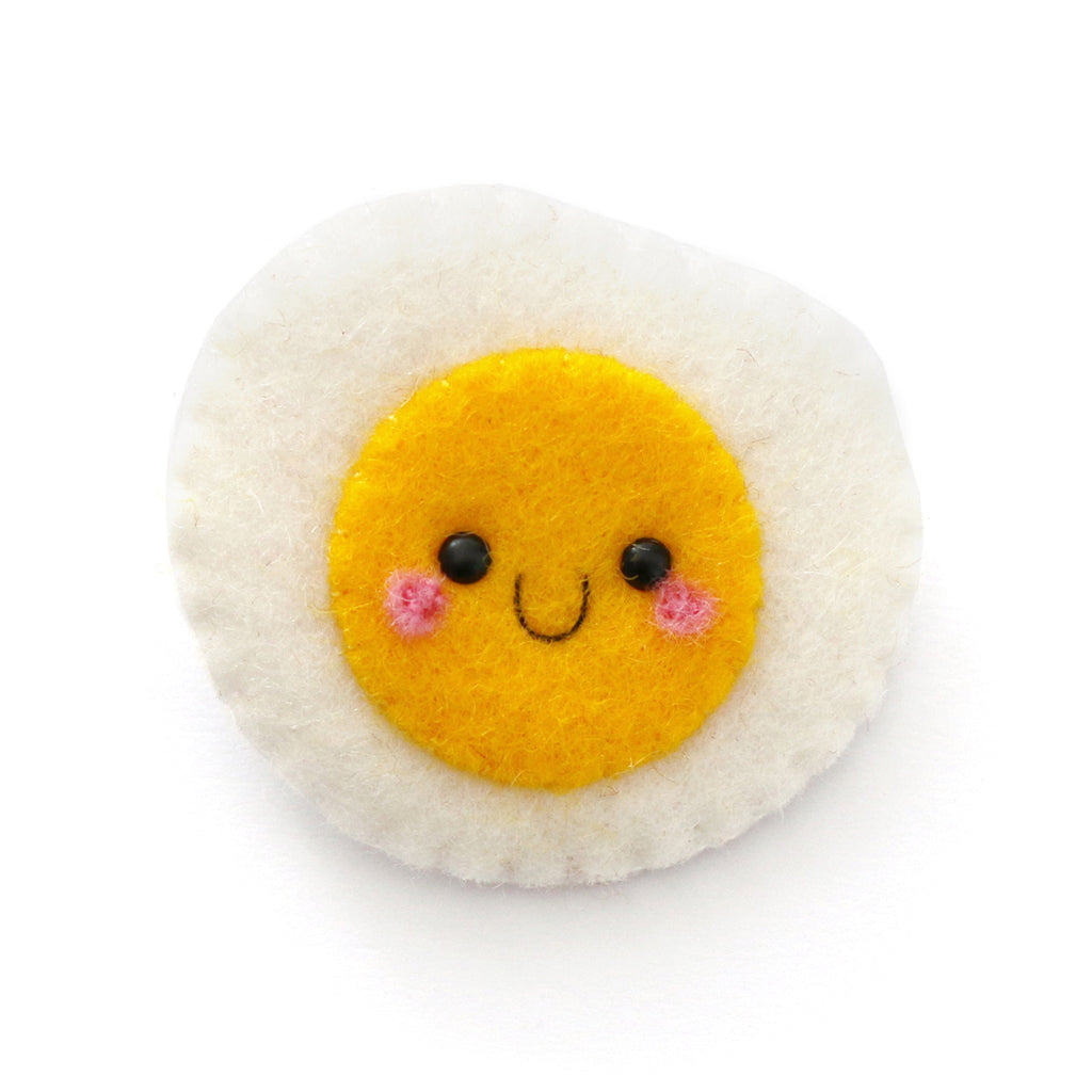 A cute, handcrafted felt egg-shaped pin with a yellow smiling face, pink cheeks, and black eyes, set against a plain white background.