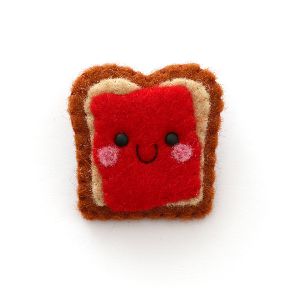 A cute felt pin shaped like a slice of toast with red jam and a smiling face, set against a plain white background.