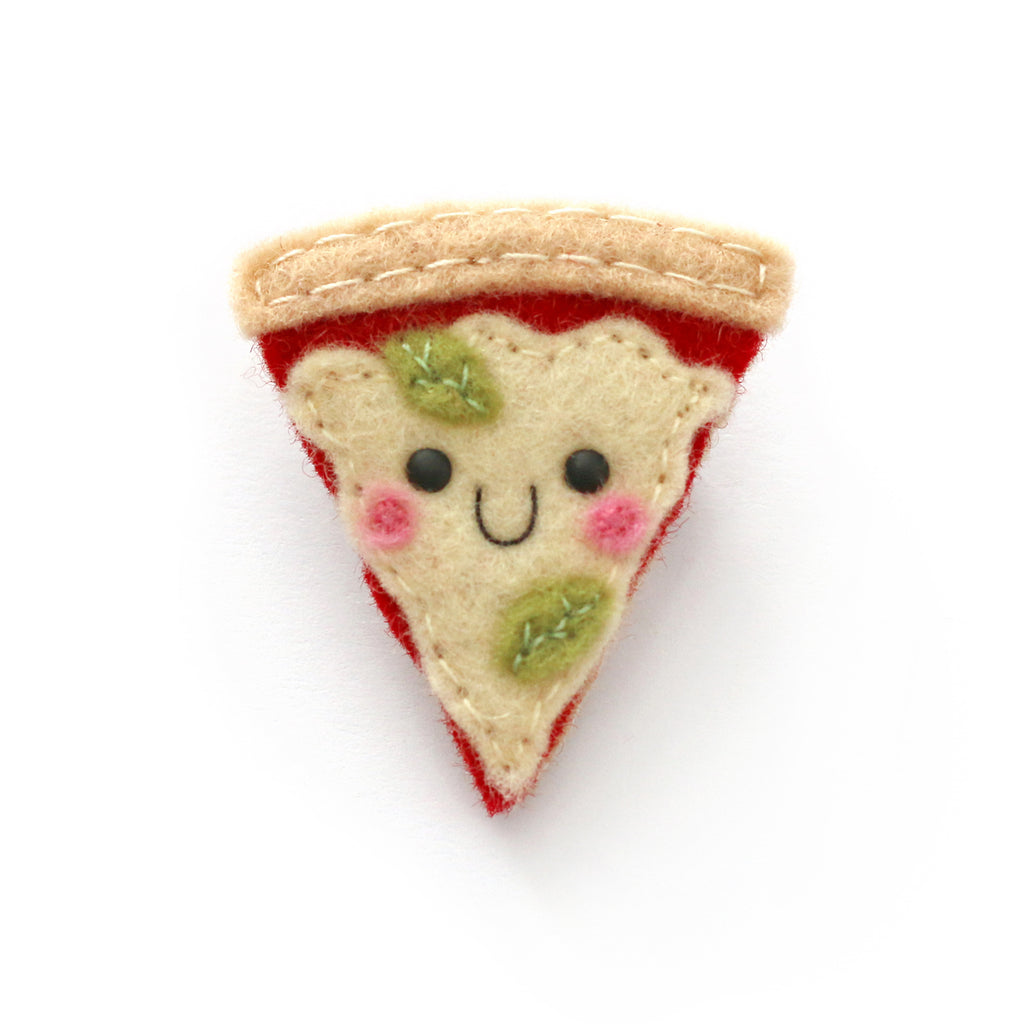 A cute, handcrafted felt pizza slice pin badge accessory with a smiling face, embroidered green basil leaves, pink cheeks, and a beige crust, shown on a plain white background.