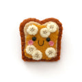 A cute, hand-felted pin shaped like a slice of toast topped with banana slices and peanut butter, on a plain white background.