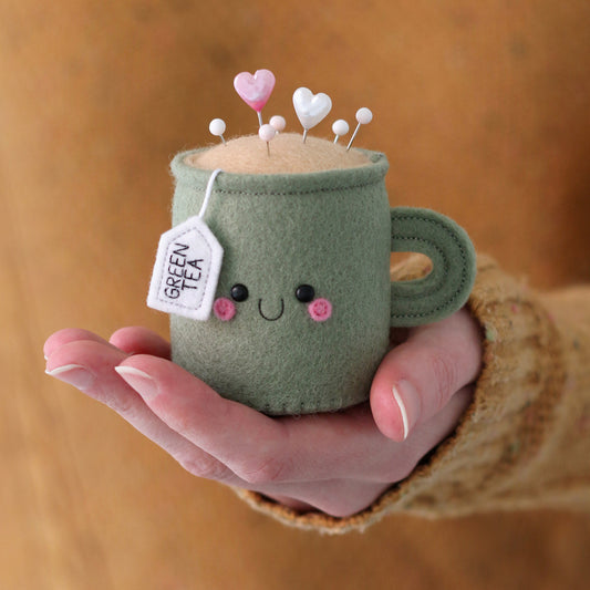 Hand holding a green felt mug with a smiling face, heart-shaped pins, and a 'Green Tea' tag. Handmade by hannahdoodle.
