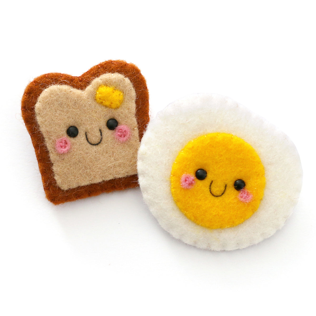 Two cute felt accessories on a white background: a toast-shaped piece with a smiling face and yellow butter detail, and a round fried egg design with a yellow yolk, and pink cheeks.