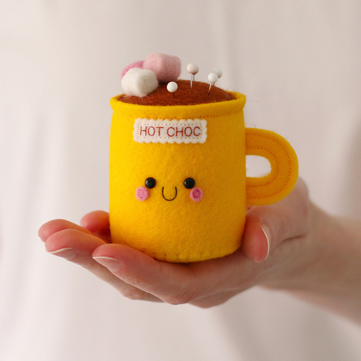 A hand holding a yellow felt hot chocolate mug pincushion with a brown hot chocolate top, pink and white marshmallows, and a smiling face on the front.