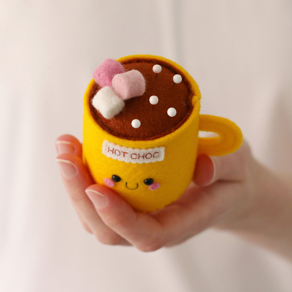 A hand holding a yellow felt hot chocolate mug pincushion with a brown hot chocolate top, pink and white marshmallows, and a smiling face on the front.