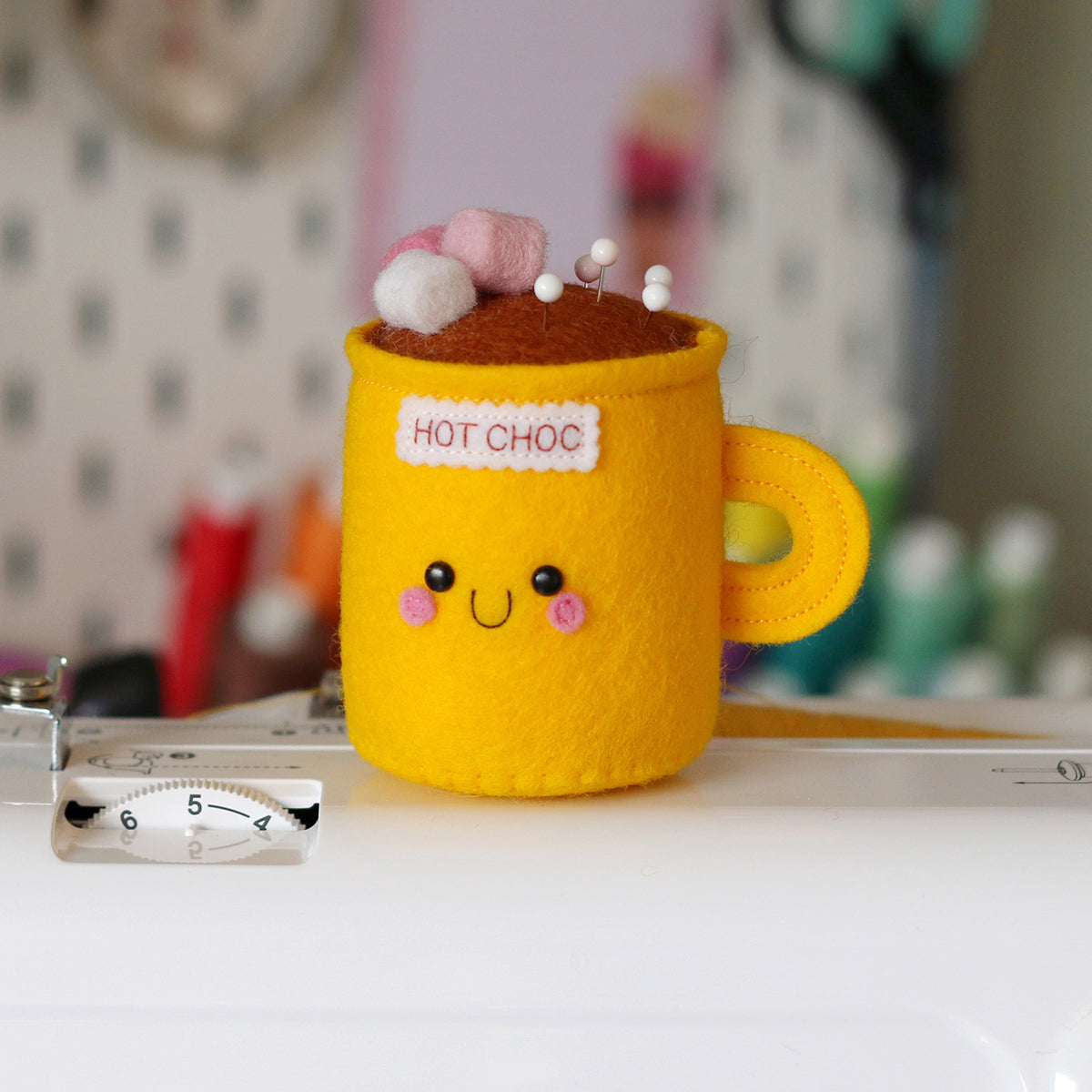 A yellow pincushion made using felt, shaped like a mug of hot chocolate, sitting on top of a sewing machine. The mug has pink and white marshmallows on top and a smiley face, with the text 'HOT CHOC' embroidered on the front. It has sewing pins placed in the top.