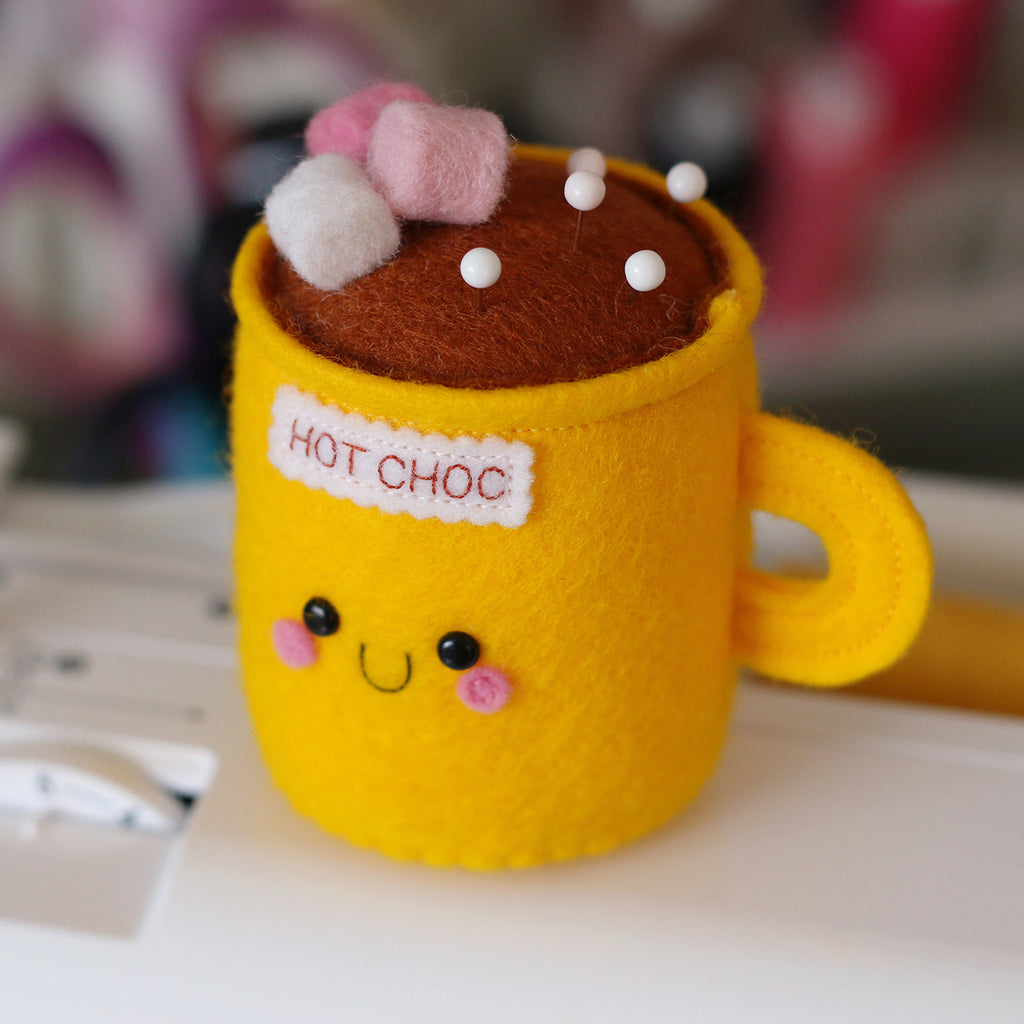 A yellow pincushion made using felt, shaped like a mug of hot chocolate. The mug has pink and white marshmallows on top and a smiley face, with the text 'HOT CHOC' embroidered on the front. It has sewing pins placed in the top.